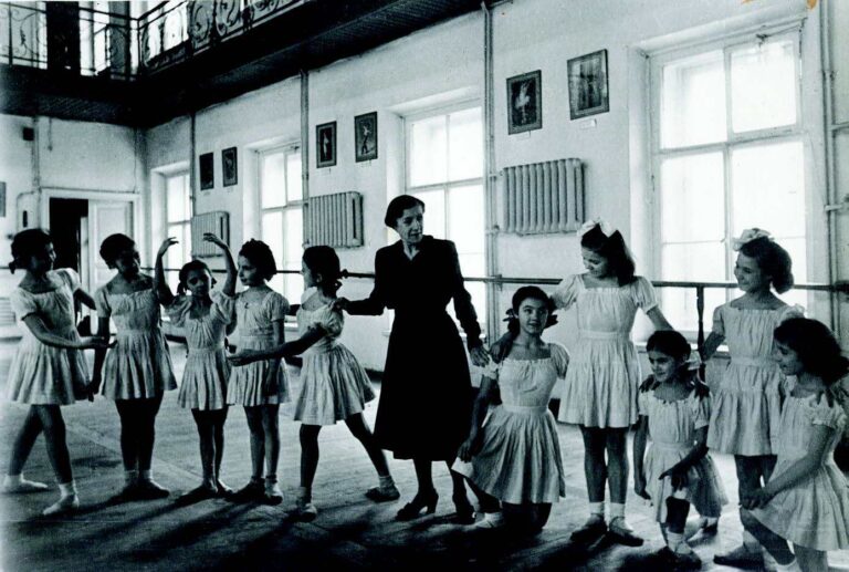 A black and white image of a dance teacher in a long black dress with young students in old-fashioned dancewear.