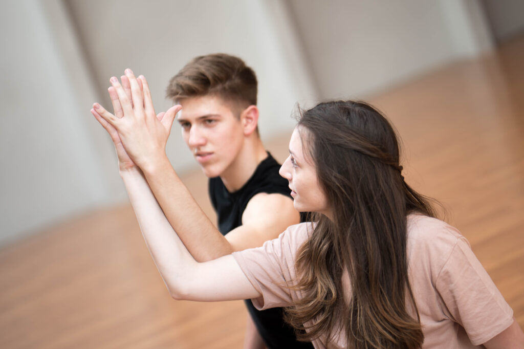 A male and female dancer, both white, touch palms of the hands with arms intertwined.