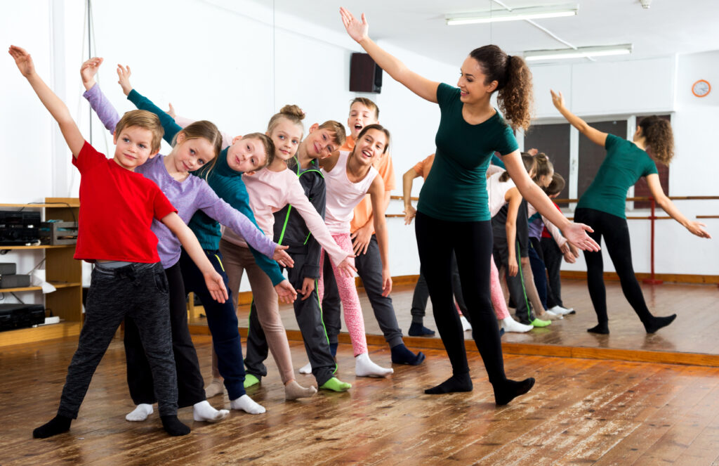 A group of children stands in a row, arms outstretched, next to a white dance teacher who is mimicking their pose.