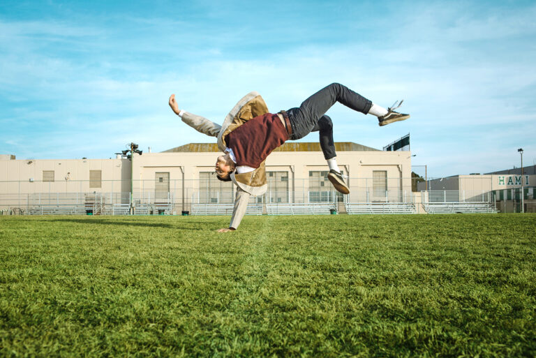 A dancer balances on one hand, seeming to float against a backdrop of grass, building, and sky.