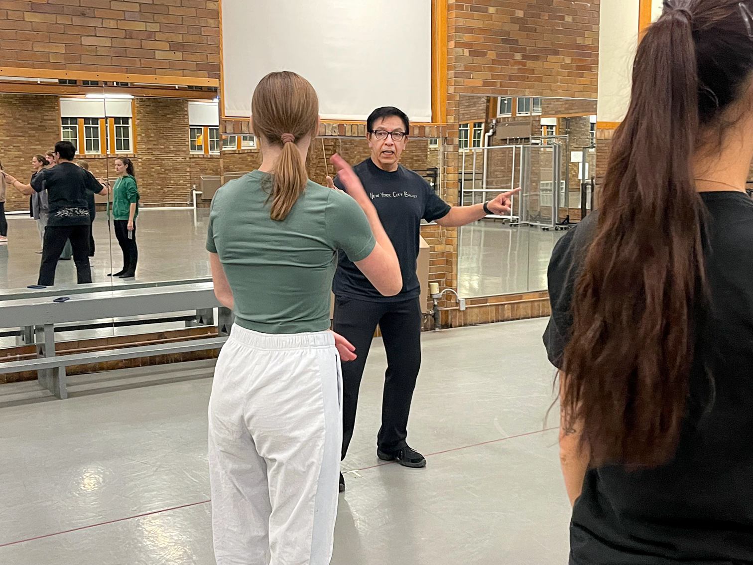 A man in a black t-shirt and black pants leads a rehearsal in a dance studio. Several dancers wearing rehearsal attire are also visible.