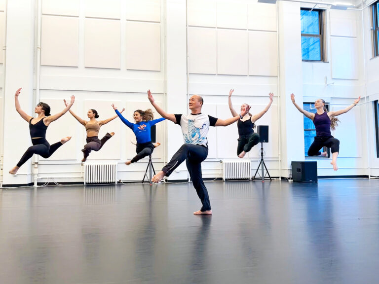 An Asian man wearing black pants and a white t-shirt stands on one leg with arms outstretched. Behind him, a group of dancers leaps with both knees bent and arms mirroring the teacher's.