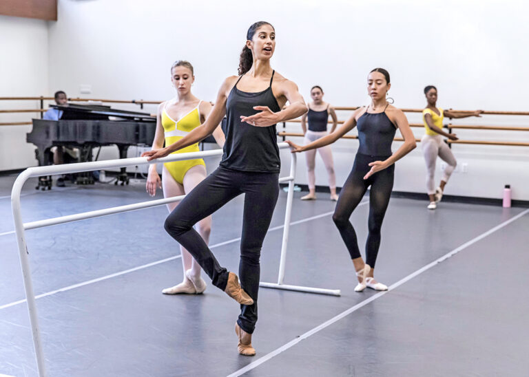 A tall, thin Black woman wearing a black tank top and leggings stands at a ballet barre, demonstrating a ballet movement to a class of dancers.
