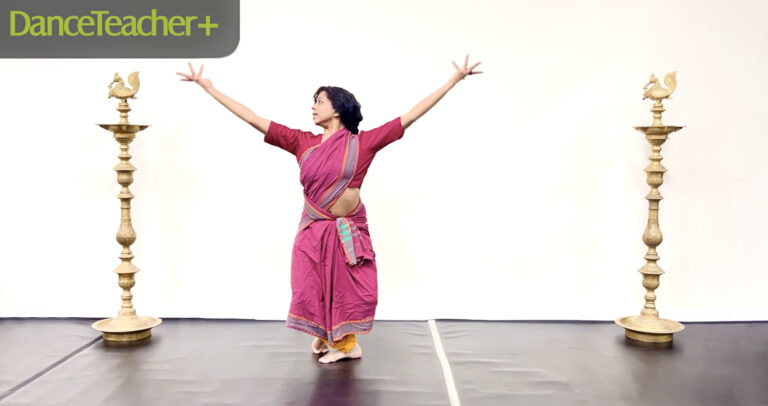 A woman in bright pink classical Indian attire is in a dance studio, arms outstretched and face tilted up toward her right hand.