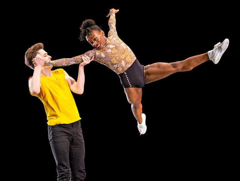 A Black dancer wearing a gold top and black shorts and white sneakers is lifted into the air, limbs outstretched, by a white dancer wearing a yellow t-shirt and black pants. Both are smiling.