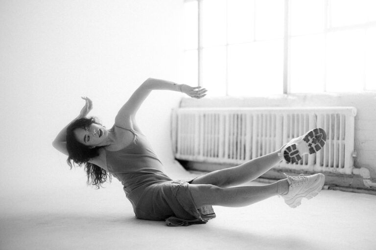 A white woman with brown hair is balanced on her pelvis, legs extended forward and arms floating upward. One arm is behind her head.
