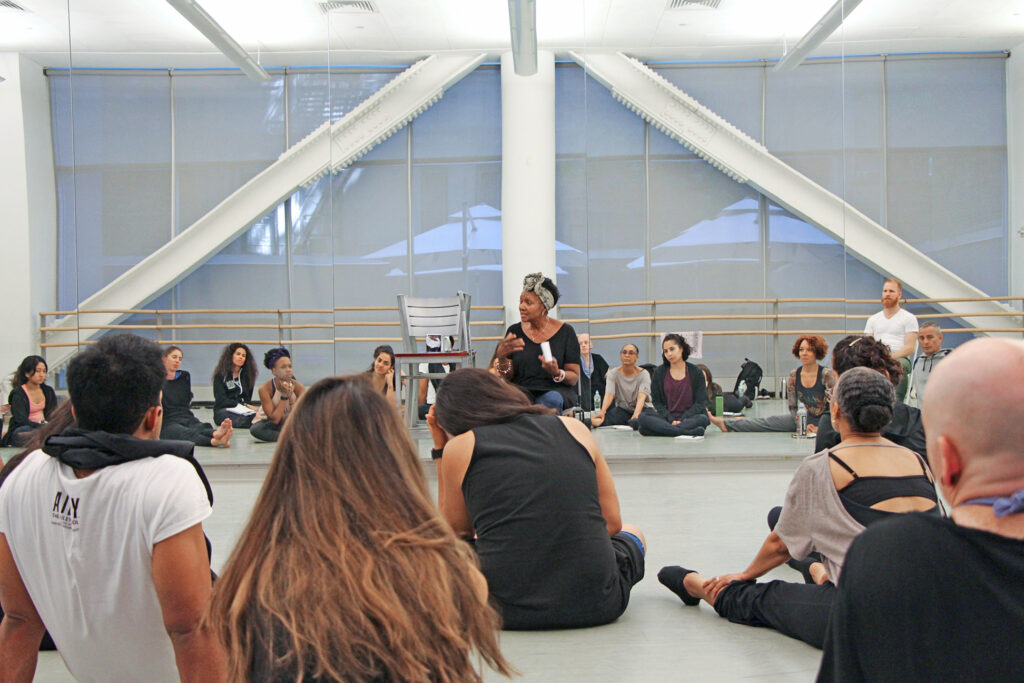 A Black woman leads a seminar with students sitting on the floor of a brightly lit dance studio.