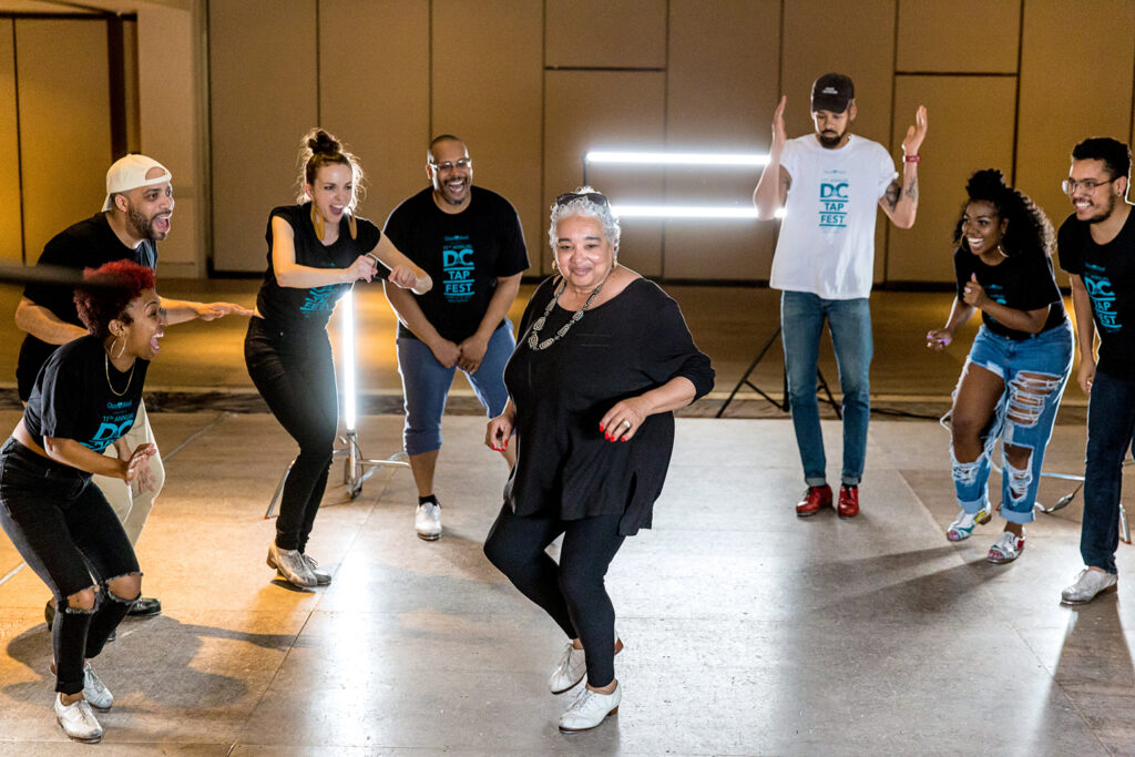 A Black woman with short white hair, wearing all black, tap dances at the center of a circle of diverse younger dancers, all of whom are cheering and clapping for her.