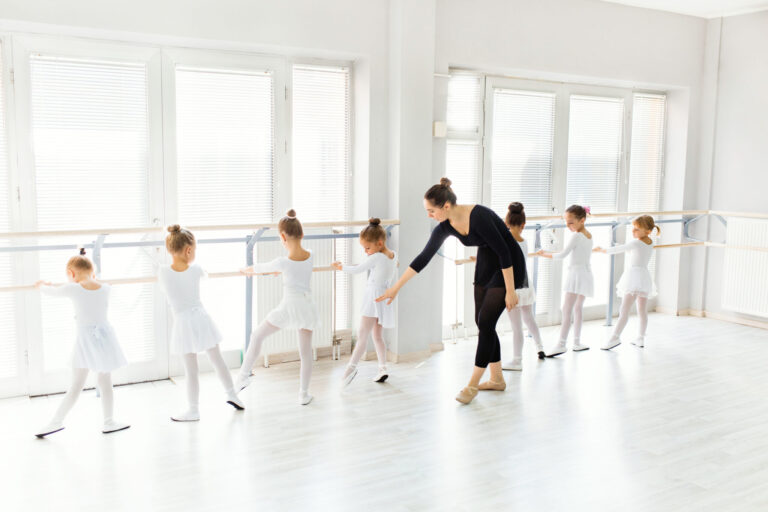 A white dance teacher in a black long-sleeved leotard, black skirt, and black leggings teaches a group of young girls at a ballet barre, all of whom wear white skirted leotards and pink tights.