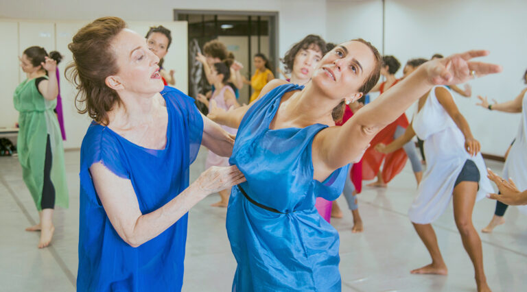 A white dance teacher wearing a bright blue tunic coaches a younger white dancer in a lighter blue tunic in an expansive arm movement.