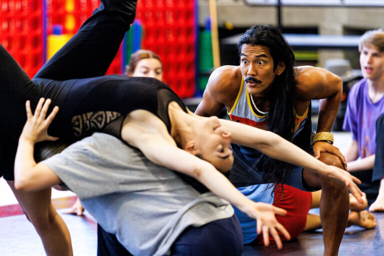 A tan man with long dark hair intently watches two dances partner each other, one in a gray t-shirt lifting another in a black tank top onto their back.