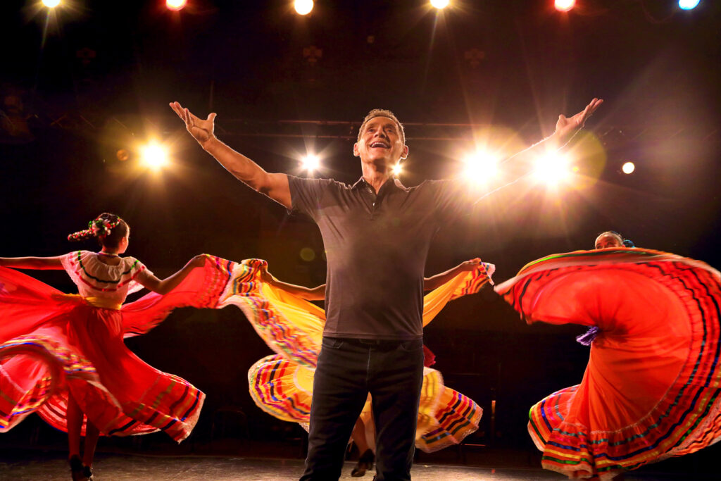 A Latino man stands center stage with arms outstretched. Behind him, dancers wearing traditional southwestern costumes twirls in bright, full red and yellow skirts. The background is black with bright spotlights.