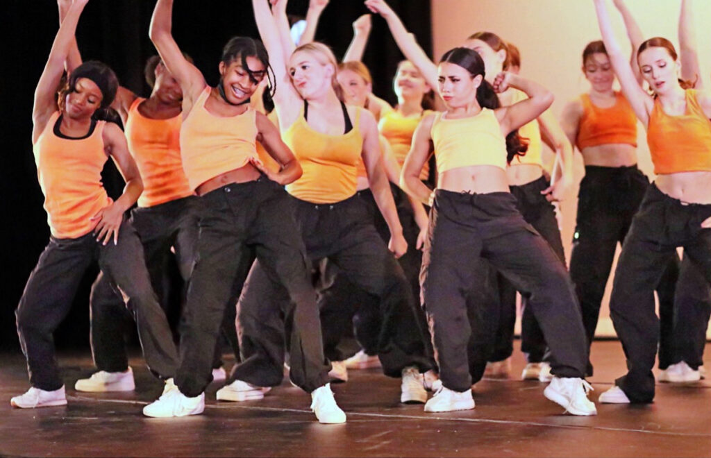 A diverse group of teenaged dancers in black pants and orange and yellow tank tops move together onstage.