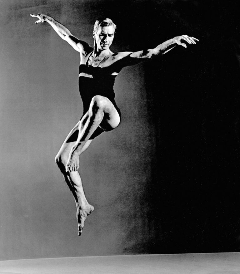A black and white photo of a male dancer jumping with knees bent and arms extended to the side above shoulder height.