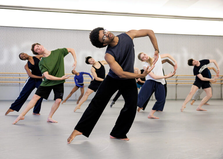 A group of Juilliard students in rehearsal for Spring Dances 2025. They all have their right foot extended to the side and arms in a circular shape learning towards the right.