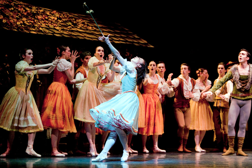 A busy crowd scene from the ballet Giselle, in which the lead ballerina, in white, is dramatically going mad while male and female ballet dancers gasp in the background.