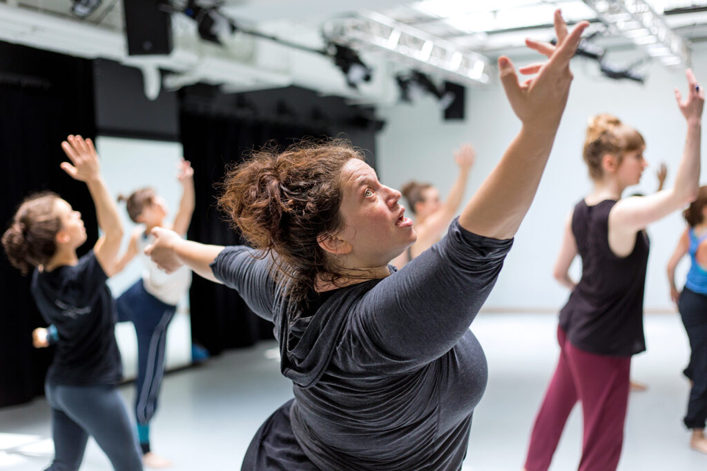Alexandra Beller wearing a grey top, arms outstretched and looking upward.
