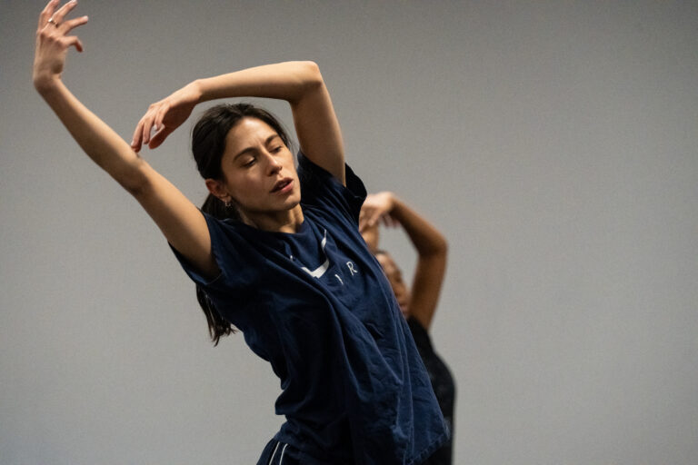 Meagan Cubides wearing a navy tshirt and arms held in a 5th position upright leaning toward the right.