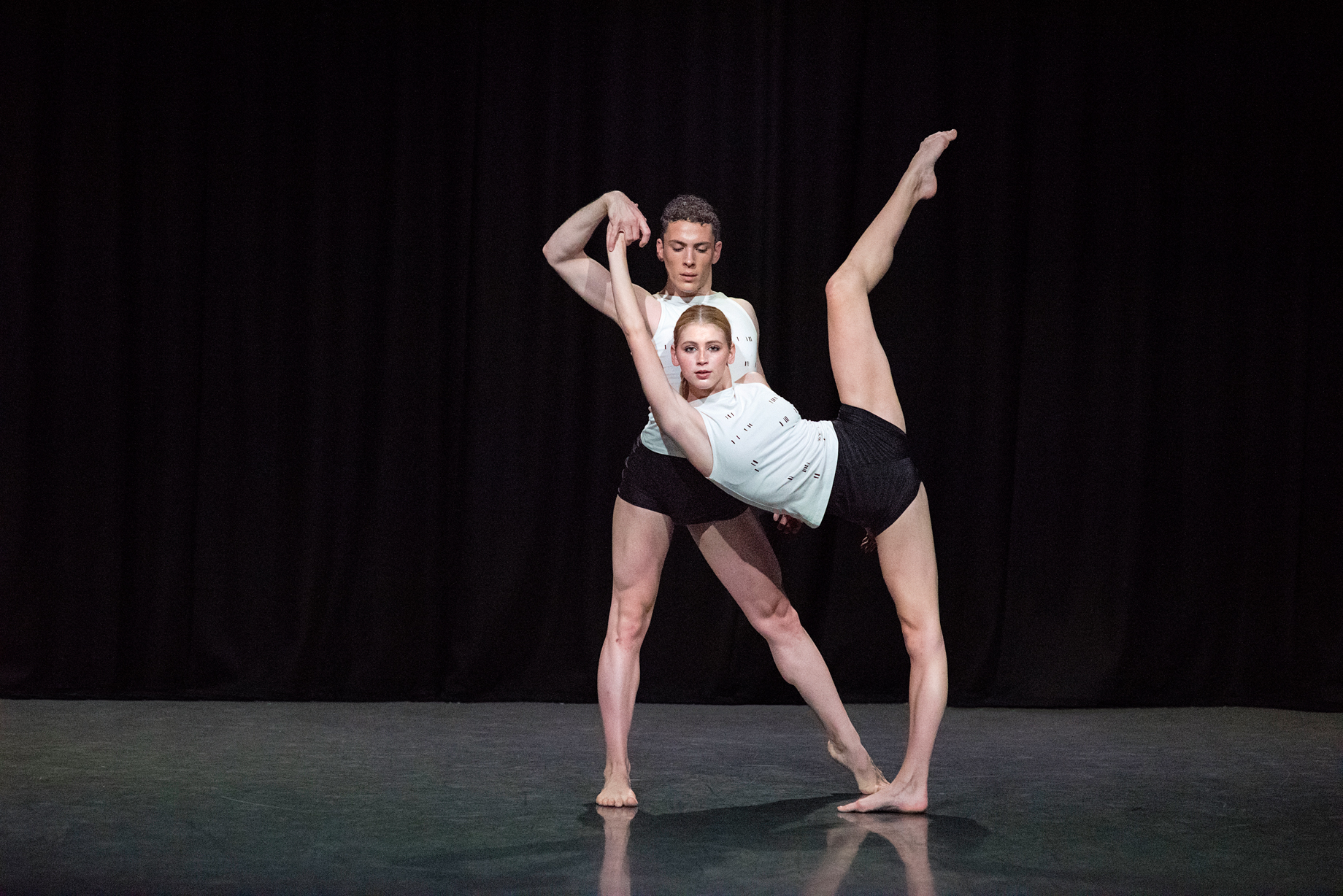 Two dancers in white tshirts and black shorts perform on stage. The female dancer in front has her left leg extended in the air bent from the knee while the male dancer holds her other hand.