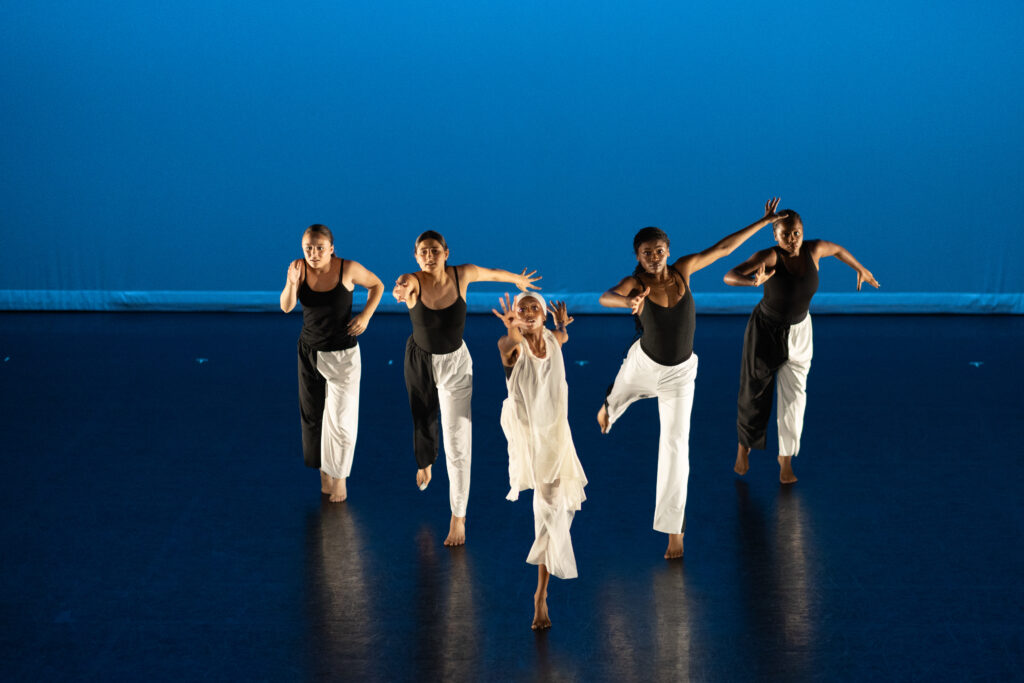Young dancers performing on stage. Four are dressed in black tops and white pants while the center dancer is dressed in all white. They perform against a blue-lit background.