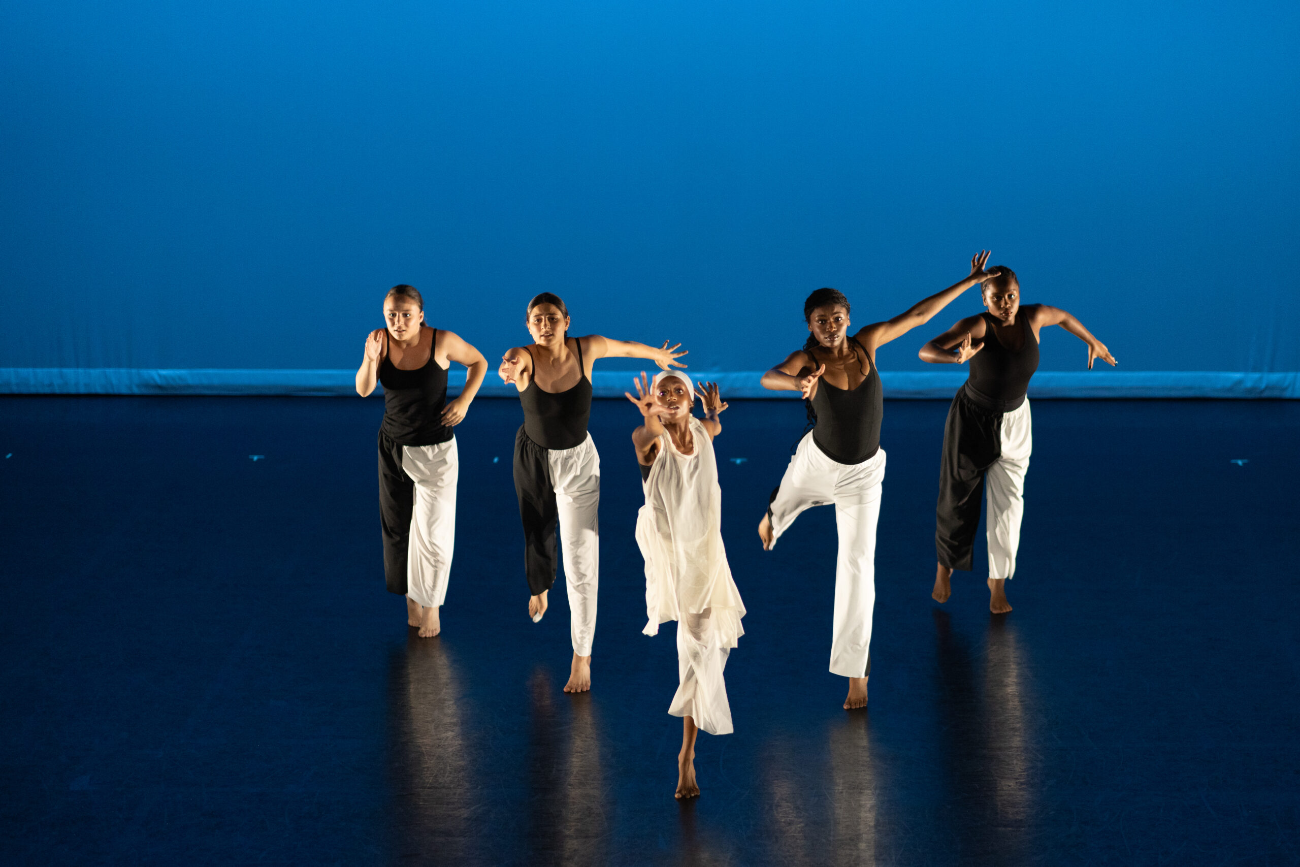 Young dancers performing on stage. Four are dressed in black tops and white pants while the center dancer is dressed in all white. They perform against a blue-lit background.