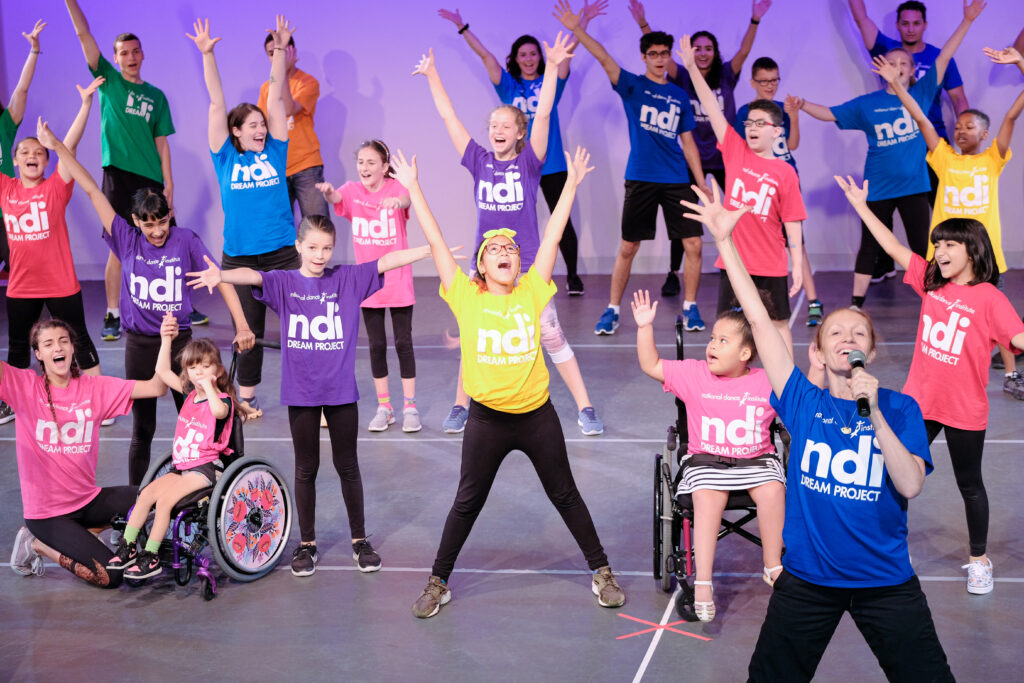 Dancers in various color tshirts with NDI written on them perform on stage.