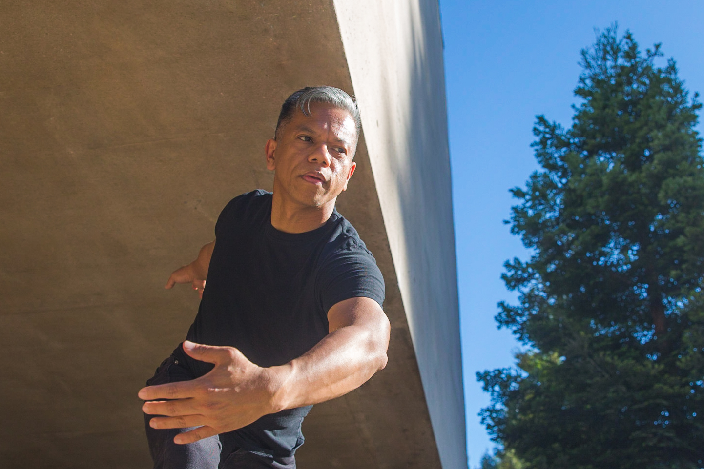 Gerald Casel dances in front of a concrete building near a redwood tree.