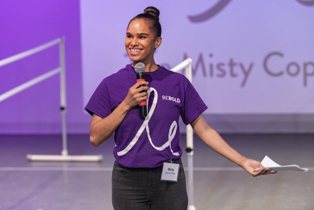 Misty Copeland holding a microphone wearing a purple tshirt with "BeBold" written on it and black jeans.