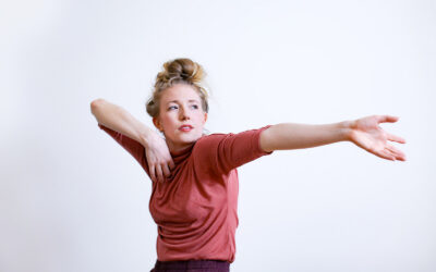 Annie Rigney in a dark rose tshirt with one arm outstretched and the other folded against her chest. She looks sideways.