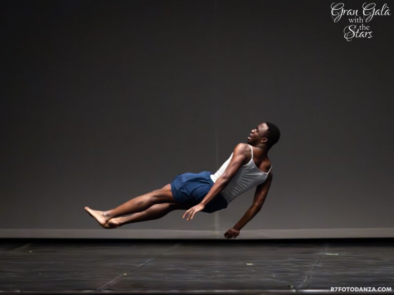 Nnamdi Nwagwu weaing a white vest and blue shorts suspended in a sideways falling pose on stage.