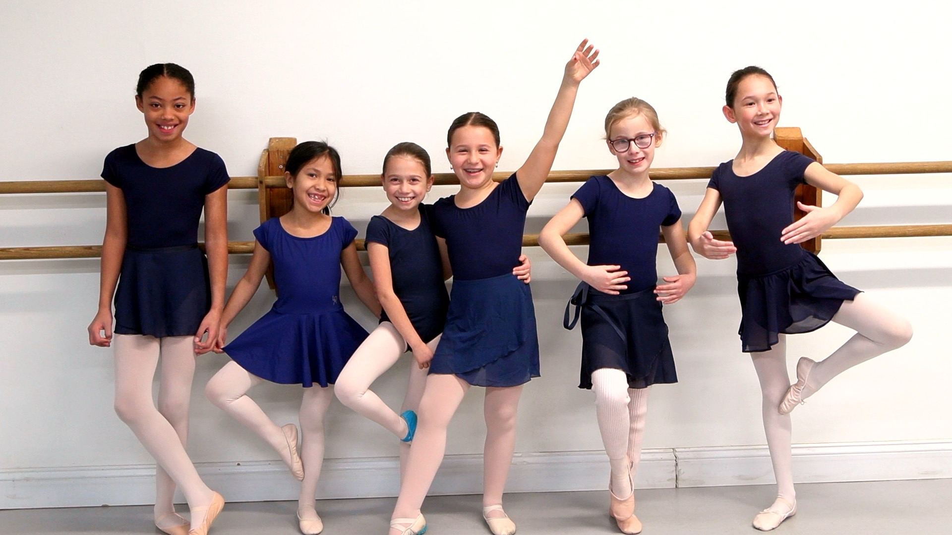 A group of six young ballet dancers/students at the barre dressed in blue leotards and skirts, pink tights, and ballet slippers.