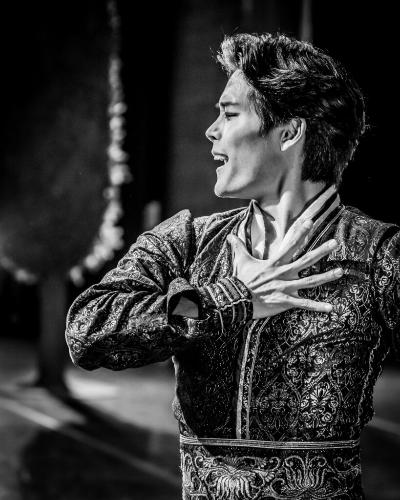 Black and white image of Choi pictured in an embellished costume from the waist up. He holds on hand to his chest.