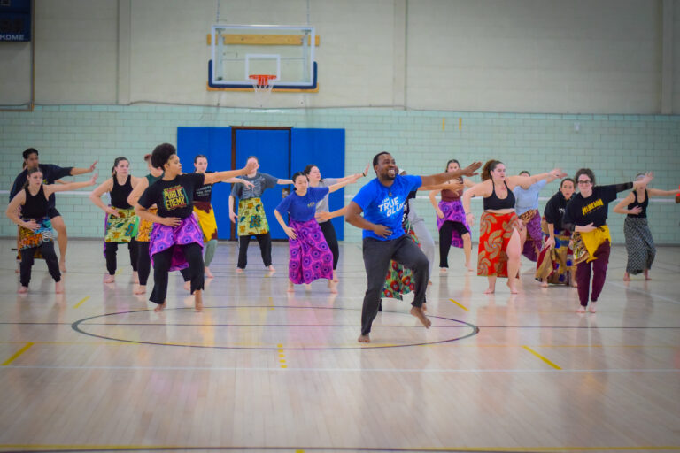 Dance students rehearsing in a basketball court wearing colorful wrap around skirts and tshirts following an instructor who is dressed in similar clothing.
