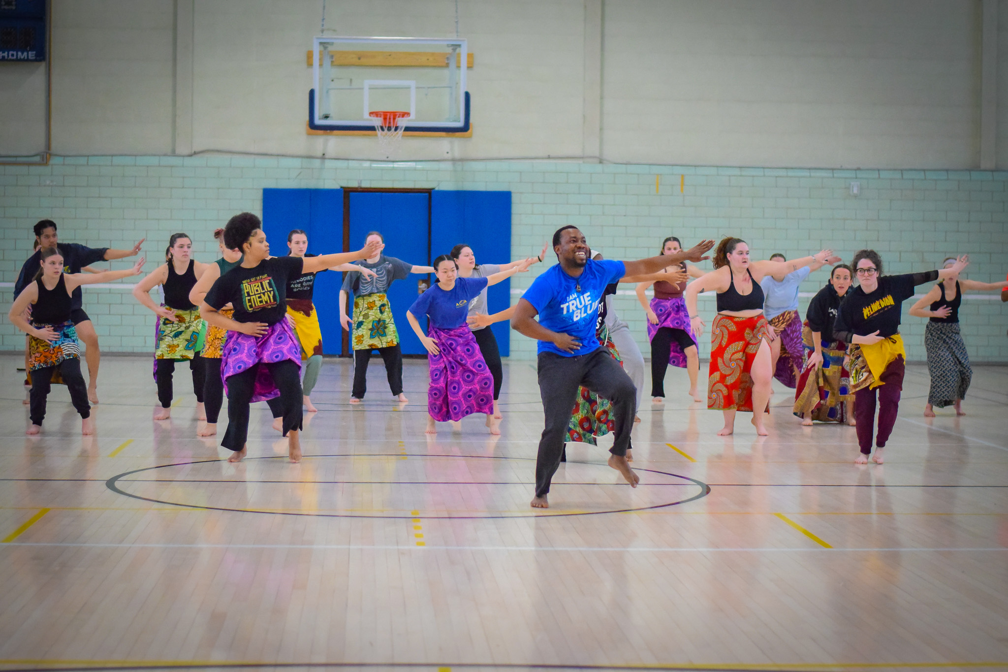 Dance students rehearsing in a basketball court wearing colorful wrap around skirts and tshirts following an instructor who is dressed in similar clothing.