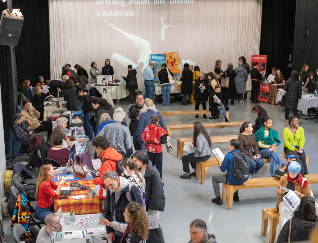 A group of attendees at a dance book fair. Benches are lined up at the middle of the room with booths to the side.