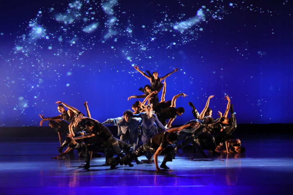A group of dancers dressed in black/dark blue costumes performing on stage.