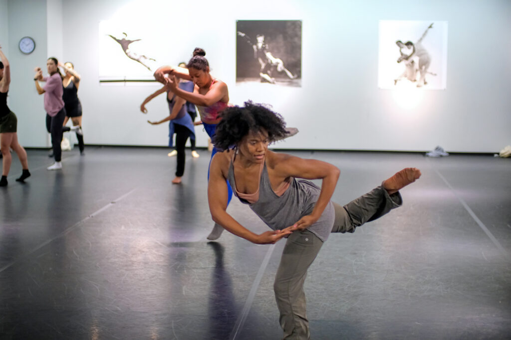 Dancer in grey tank and sweatpants turns to the side with arms in a circle held below her waist.
