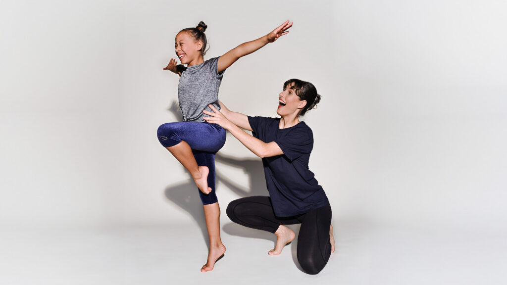 A young dancers balances on one leg with her arms outstretched while an instructor kneels beside her, supporting her at the waist.