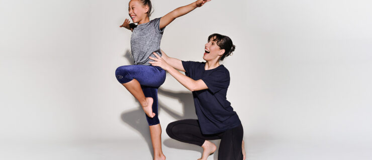 A young dancers balances on one leg with her arms outstretched while an instructor kneels beside her, supporting her at the waist.