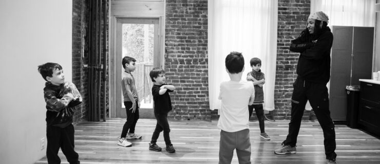 Black and white photo of a dance instructor leading a group of young children practicing a cross-armed movement in a studio with brick walls and wood floors.