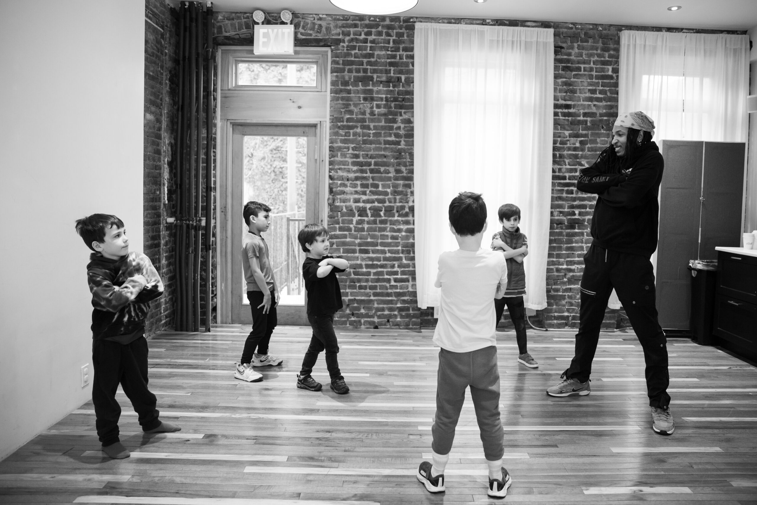 Black and white photo of a dance instructor leading a group of young children practicing a cross-armed movement in a studio with brick walls and wood floors.