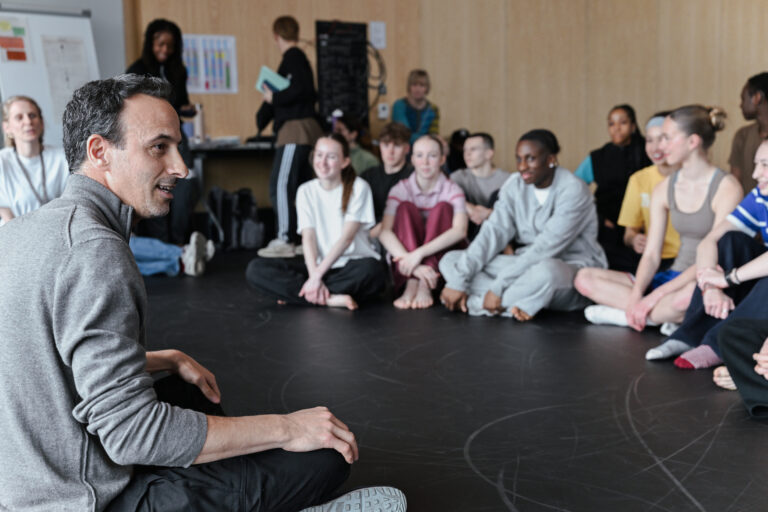 Hofesh Shechter sits on the floor of a studio, speaking to a seated circle of young dancers in warm-ups.