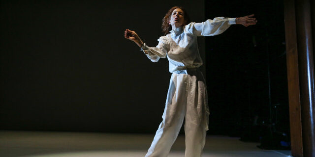 A woman in baggy white clothing dances under a spotlight in a darkly lit room.