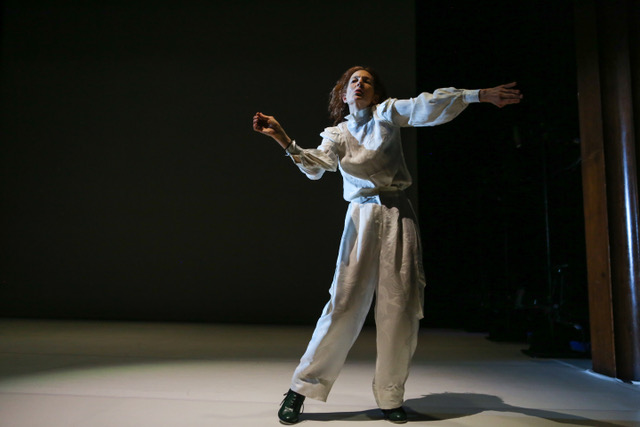 A woman in baggy white clothing dances under a spotlight in a darkly lit room.