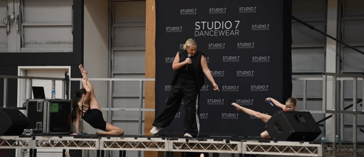 On a dance convention stage, a girl on the far left is sitting on her left leg with her right leg extended. In the middle, Shannon Atkins points at a dancer on the far right, who is moving into a similar leg extension.