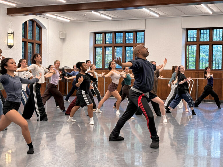 A group of students in a dance studio imitate the movement of a faculty member.