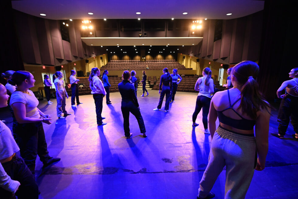 From an upstage perspective, a group of dancers stands on a stage underneath purple lighting listening to a faculty member. 
