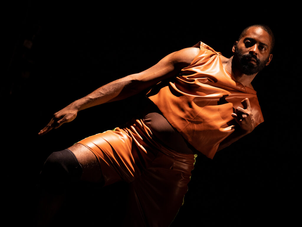 Image of dance artist Jerron Herman clothed in a metallic orange outfit holding a pose sideways against a black background. He holds two fingers up.
