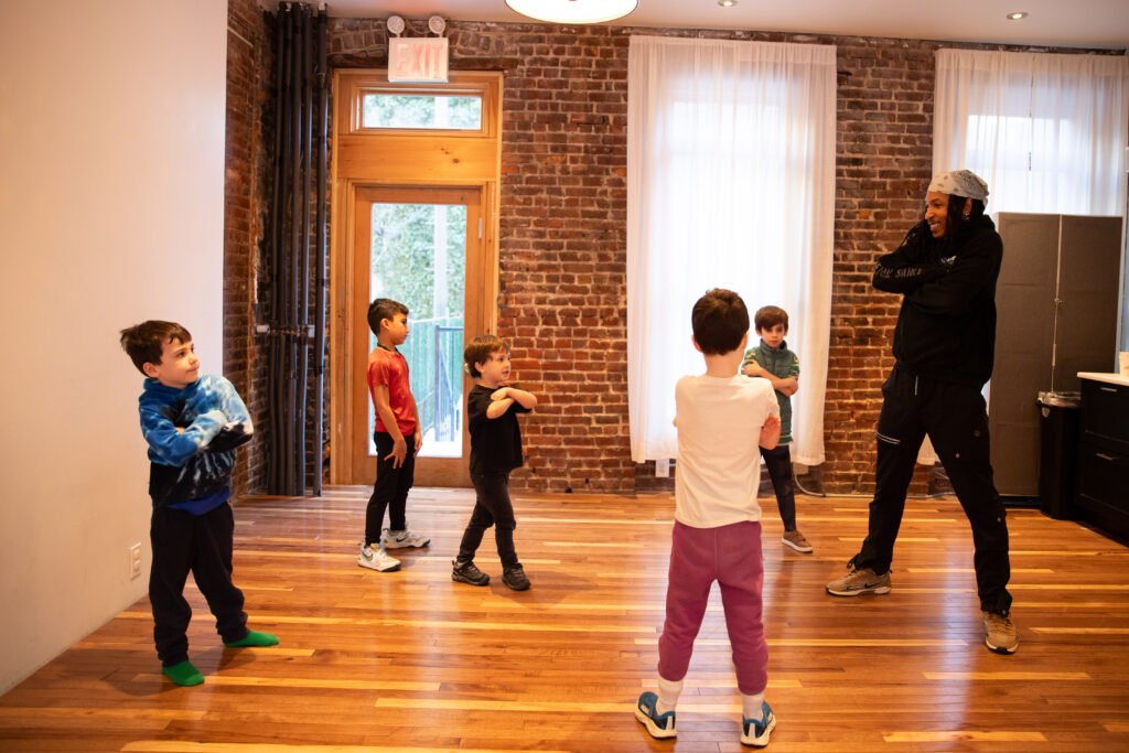 Photo of a dance instructor leading a group of young children practicing a cross-armed movement in a studio with brick walls and wood floors.