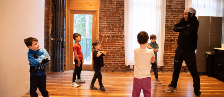 Photo of a dance instructor leading a group of young children practicing a cross-armed movement in a studio with brick walls and wood floors.